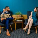 A man and woman engaged in conversation, sitting in stylish armchairs against a blue wall with a wooden side table and potted plants.